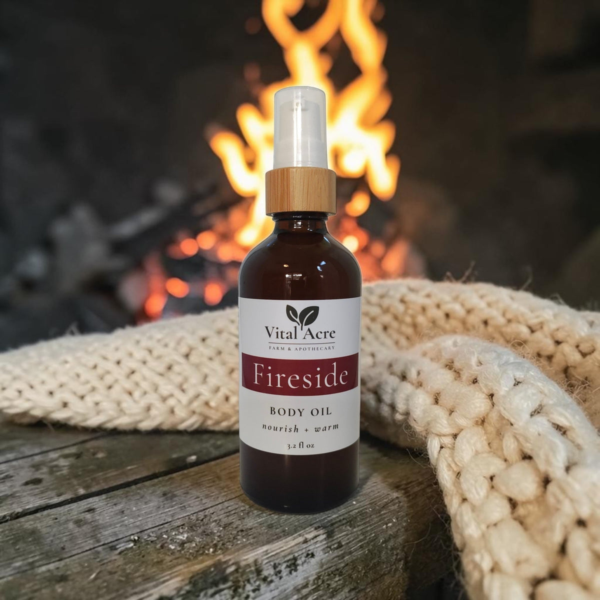 A bottle of Vital Acre Fireside body oil stands on a rustic wooden surface with a knitted cream blanket draped around it and a blurred fireplace in the background.