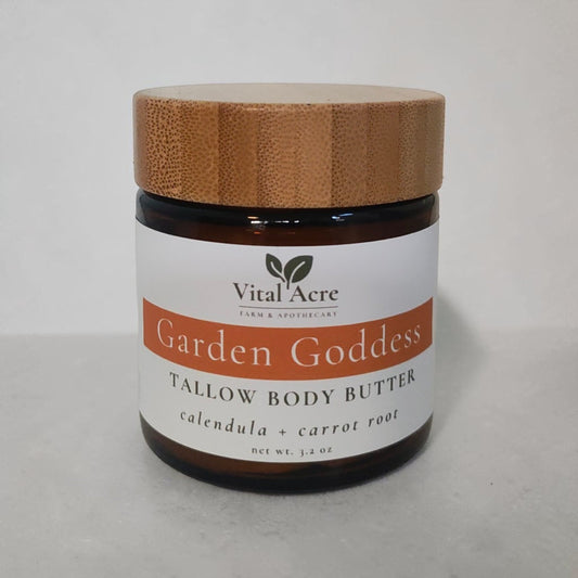A jar of Vital Acre Garden Goddess Tallow Body Butter with a white label and a light brown bamboo-style lid, set against a neutral light gray background.