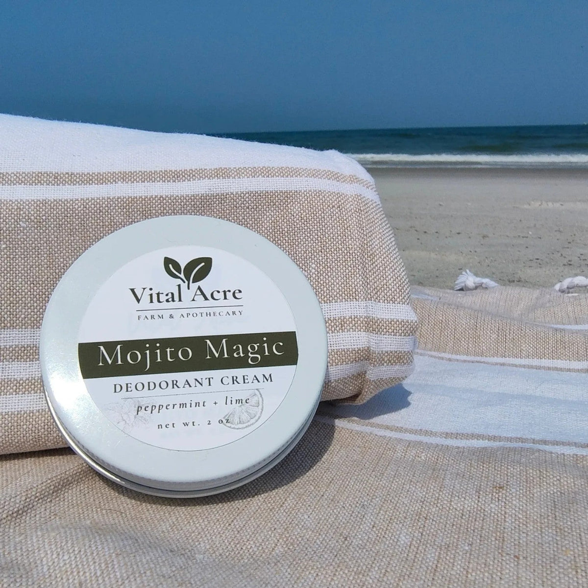 Vital Acre Farm & Apothecary Mojito Magic aluminum-free deodorant cream tin on a striped beach towel by the ocean.